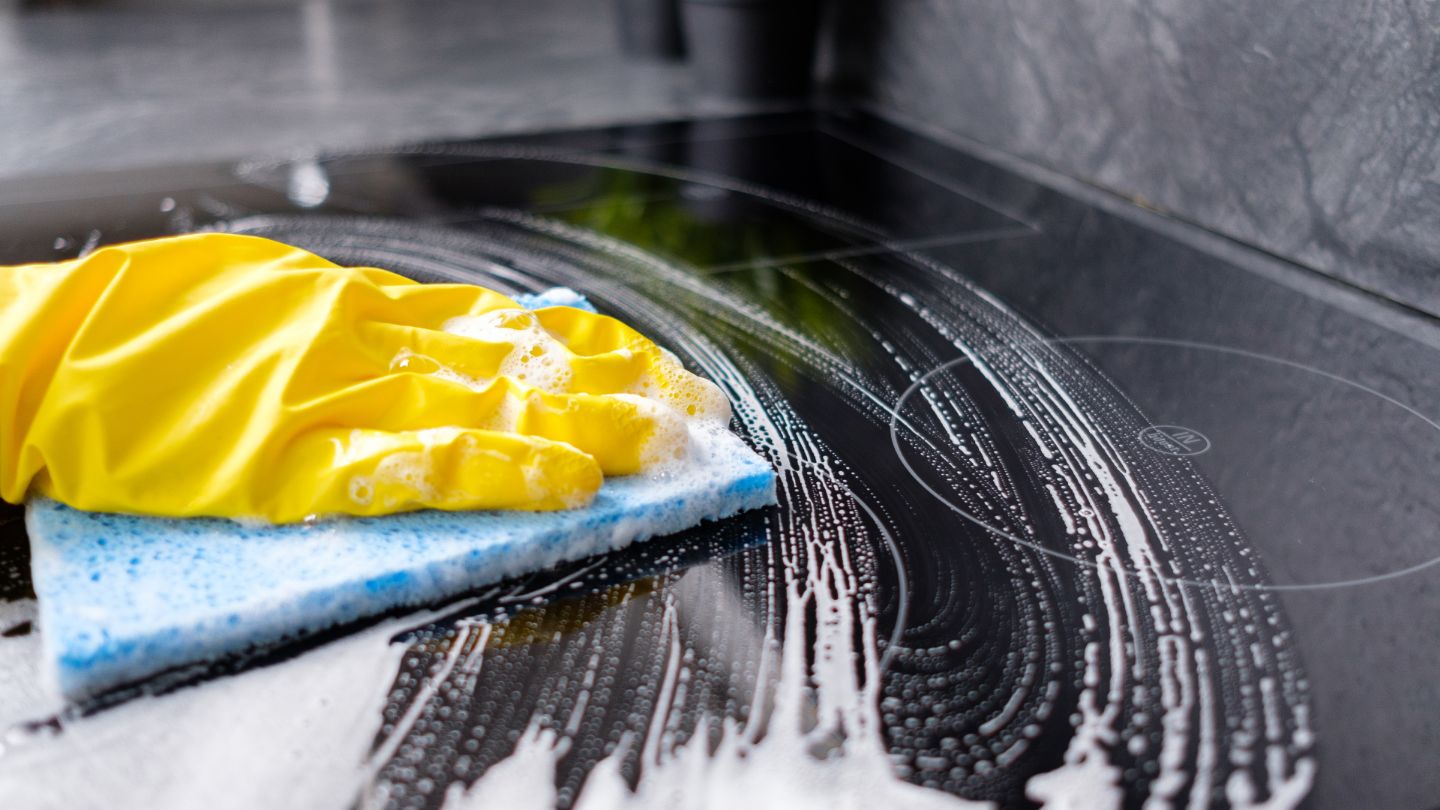 Professional deep cleaning of kitchen stovetop with yellow gloves and cleaning sponge creating suds on black surface